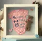 Preview: Stickdatei Set Hello Baby (div. Größen), Foto zeigt tiefen Bilderrahmen mit Babybody und Stoffteddy und Herzen als Schmuckbeigabe. Body rosa mit weißen Punkten. In Blautönen Bestickung mit Maßband und Schrift "52 cm", Hantel und Schrift "3380 g", Wecker u