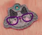 Preview: Stickdatei Set I'm Happy Cat Doodle Applikation inkl. ITH Anhänger, Foto zeigt Anhänger auf felliger Unterlage. Katzenkopf, oben stoffgeöst, in entsprechendem Umriss. Farben hell- und dunkelgrau, lila Brille.