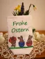 Preview: Stickdatei Set Osterhasen ITH Lichtbeutel / Utensilos, Foto zeigt hellen Hochkantbeutel vor Frühlingsdeko. Bunte Bestickung: 3 diverse Hasen mit Ei/Eierschale auf Wiese, darüber Schriftzug "Frohe Ostern".