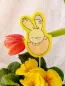 Preview: Stickdatei Set Osterhasen ITH Stabdesigns, Foto mit Designstab Blumentopf mit Blume steckend, im Hintergrund neigt sich eine Tulpe. Auf gelbem Filz gestickter Osterhase, der in halber Eierschale sitzt und sich nach unten beugt.