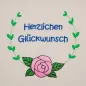Preview: Stickdatei Set florale Rahmen + Schriftzüge,  Foto mit Bestickung in grün-rosa-blau auf hellem Stoff. Von einer Rose mit Blättern führen 2 gebogene Blattzweige randgebend nach oben, dort Rahmen offen. Innen der Schriftzug "Herzlichen Glückwunsch".