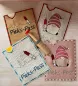 Preview: Stickdatei Set ITH Impfpass-Hüllen 24 Designs (13x18), Foto mit 4 beispielhaften Hüllen, auf Holzuntergrund liegend, darauf Holzspritze als Accessoire. 2 mal Spritzengnom; 2 mal EKG-Kurve mit Spritze. Alle zusätzlich mit Schriftzug "Pieks-Pass".