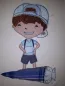 Preview: Stickdatei Set Schulkind Liam (div. Rahmengrößen), Foto von Applikation auf hellem Stoff. Braunhaariger Junge mit Basballcap, der hinter einer liegenden Schultüte steht. In Blautönen gehalten.