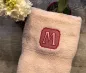 Preview: Stickdatei Set Monogramme Lotte geeignet für Frottierwaren, Foto zeigt zusammengelegtes helles Handtuch auf Holztisch, darauf anteilig Blüte und Topfpflanze. Buchstabe "M" aufgestickt, hell auf rotem Rahmen.