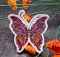 Preview: Stickdatei Schmetterling Vollstickmotiv, Foto mit Ausfertigung auf dunkelgrauem Untergrund mit hereinragenden orangen Blumen. Motiv: Schmetterling in rot-gelb-pink-schwarz.