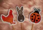 Preview: Stickdatei Set ITH Frühlings-Stabdesigns, Foto mit 3 Designstäben, auf orangem Fell liegend. Motive: Huhn (rot), Hase (braun-weiß), Marienkäfer (rot-schwarz).