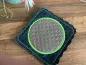 Preview: Stickdatei Set ITH Mug Rug 10. Der Ausschnitt zeigt auf einem Holzuntergrund  den Mug Rug als quadratischen dunkelgraublauen wellig geschnittenen Filz mit hellgrün abgestepptem Rand. Mittig ein wellig-blattartiges Ornament in grau, das mit dem hellgrünen