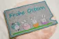 Preview: Stickdatei Set Osterhüllen ITH Geschenk- und Schokoladenhüllen + Einzelmotive. Foto, das eine Ausfertigung auf apricotfarbenem Fell liegend zeigt. Querformatige Hülle aus grauem Filz mit Schriftzug "Frohe Ostern", darunter 4 verschiedene Osterhasenmotive