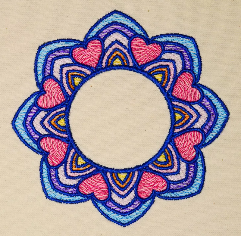 Stickdatei Set Mandalas + Split Mandala (div. Größen), Foto von Ornament auf hellem Stoff. Buntes Mandala-Ornament in blau-rot-lila-gelb-rosa aus abgerundeten Zacken und Herzen und mit kreisrundem, freiem Zentrum.