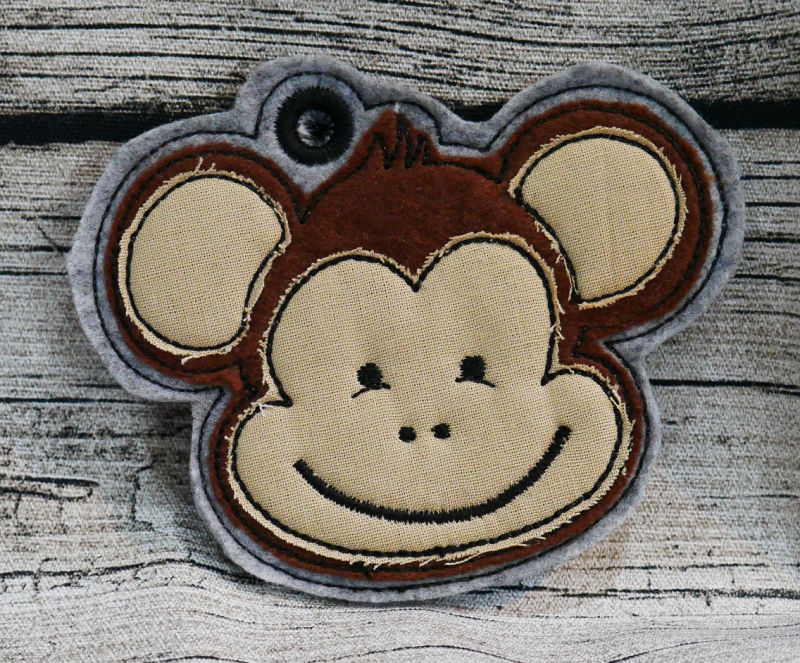 Stickdatei Affe Applikation in 5 Größen + ITH Anhänger, Foto zeigt auf Holzuntergrund liegenden garngeösten Anhänger. Auf grauem Filz lächelnder Affenkopf in braun-beige.