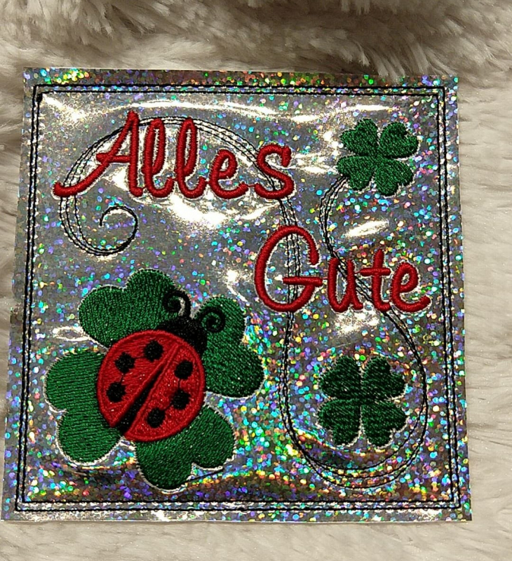 Stickdatei Set ITH Mini-Grußkarten inkl. Abwandlung zur Geschenktasche, Foto mit 1 quadratischen Ausarbeitung, präsentiert auf hellem Untergrund. Glitzerbunter Hintergrund mit bunter Bestickung Glücksklee, Marienkäfer, Schriftzug "Alles Gute".