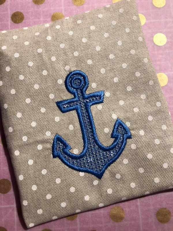 Stickdatei Set Maritime Handtuch Motive Embossed 10x10, Foto von grau-braunem Stoff mit weißen Punkten, darauf in blau gestickt ein Anker. Dieser hat eine dicke Umrandung, die Füllfläche ist gemustert.