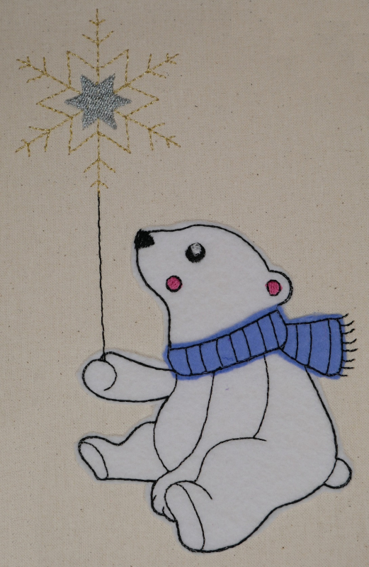 Stickdatei Set Eisbären Applikationen, Foto mit bestickter Applikationen auf hellem Stoffuntergrund. Weißer Eisbär trägt blauen Schal. In der Hand Schneeflocke.