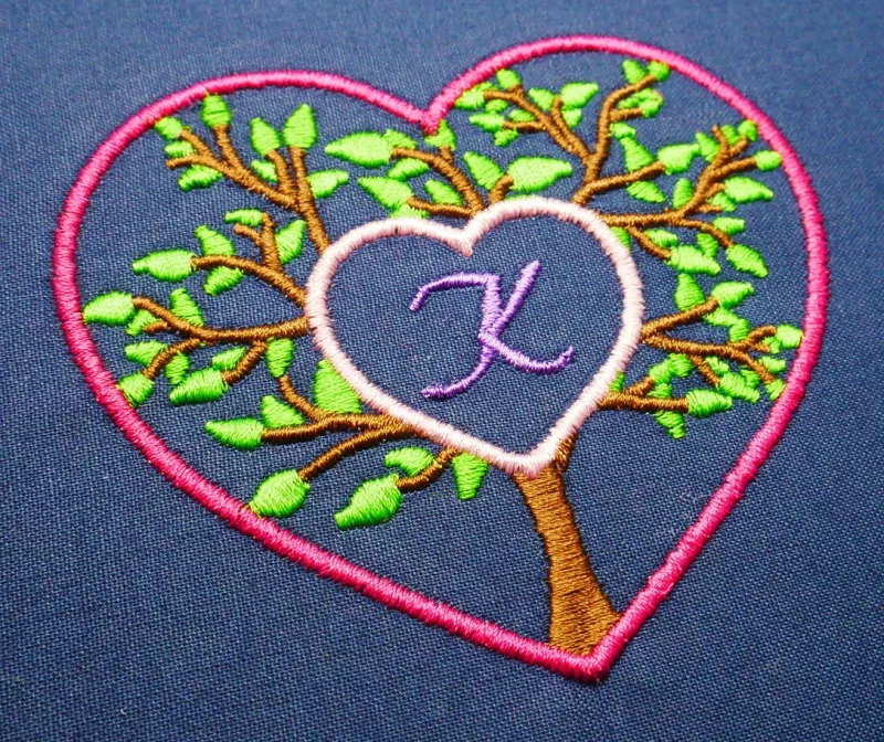 Stickdatei Set Monogramm Motiv Baum inkl. ITH Schild. Foto zeigt auf dunkelblauem Stoff pinken Herzumriss, darin ein grün-brauner Baum, in dessen Zentrum kleine Herzumrandung rosa, darin Baumstick ausgespart und mit pinkem Buchstaben "K" stattdessen.