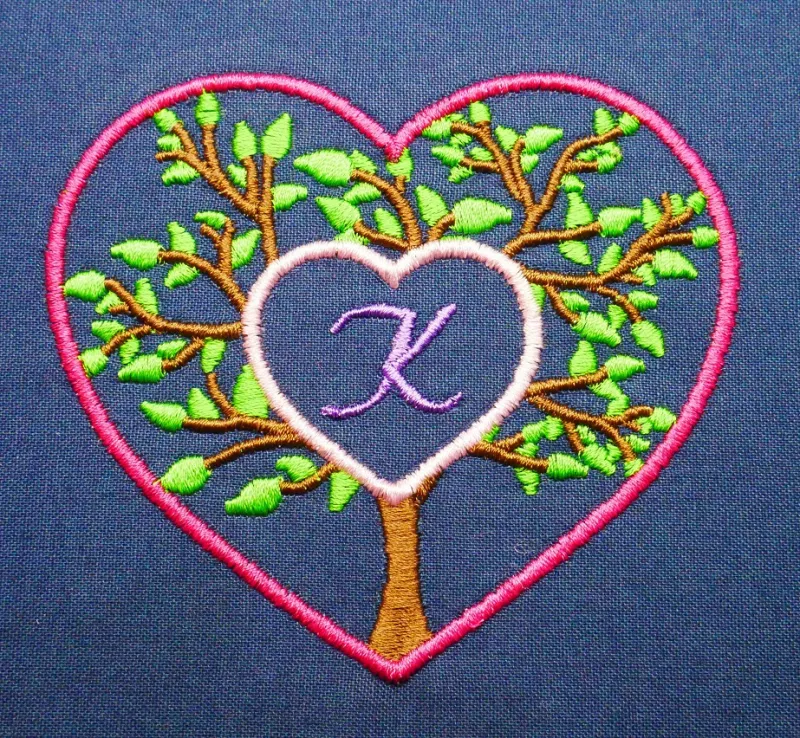 Stickdatei Set Monogramm Motiv Baum inkl. ITH Schild, Foto zeigt pinken Herzumriss auf dunkelblauem Stoff. Innerhalb des Herzumrisses Baum mit grünen Blättern, Zentrum ausgespart und gefüllt mit kleinem rosa Herzumriss, das den Buchstaben "K" zeigt.