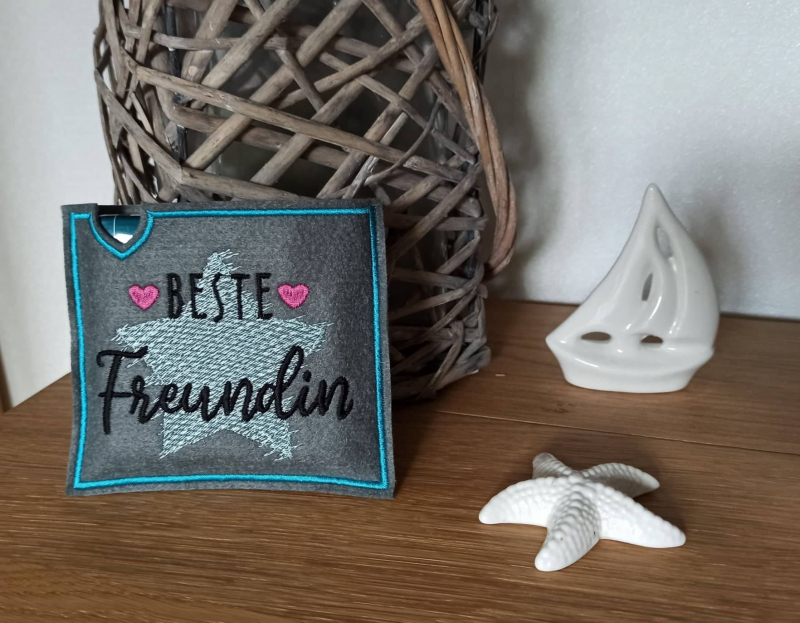 Stickdatei Set "die Besten" mit ITH Mug Rugs, Schokohüllen & Schriftzügen, Foto zeigt querformatige Hülle in dunkel. Aquamarinfarbener Stern, Schriftzug "Beste Freundin", zwei rote Herzchen.