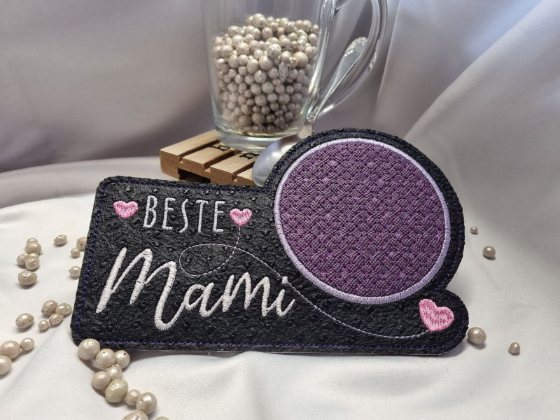 Stickdatei Set "die Besten" mit ITH Mug Rugs, Schokohüllen & Schriftzügen.
Foto zeigt schmalen Tassenteppich aus schwarzem Filz mit darüber hinausgehendem kreisrundem lila Abstellplatz. Schriftzug "Beste Mami"/Schnörkel/rosa Herzchen.