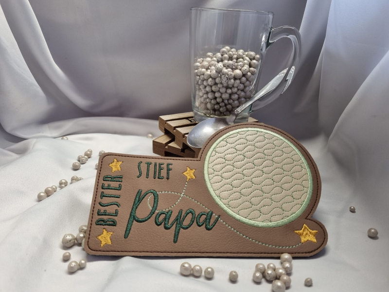 Stickdatei Set ITH Mug Rugs "die besten" inkl. Schriftzüge. Foto zeigt schmalen Tassenteppich aus braunem Kunstleder mit darüber hinausgehendem kreisrundem Abstellplatz mit beige-grünem Muster darin. Bestickung: "Bester Stiefpapa" mit Schnörkel und 4 gelb