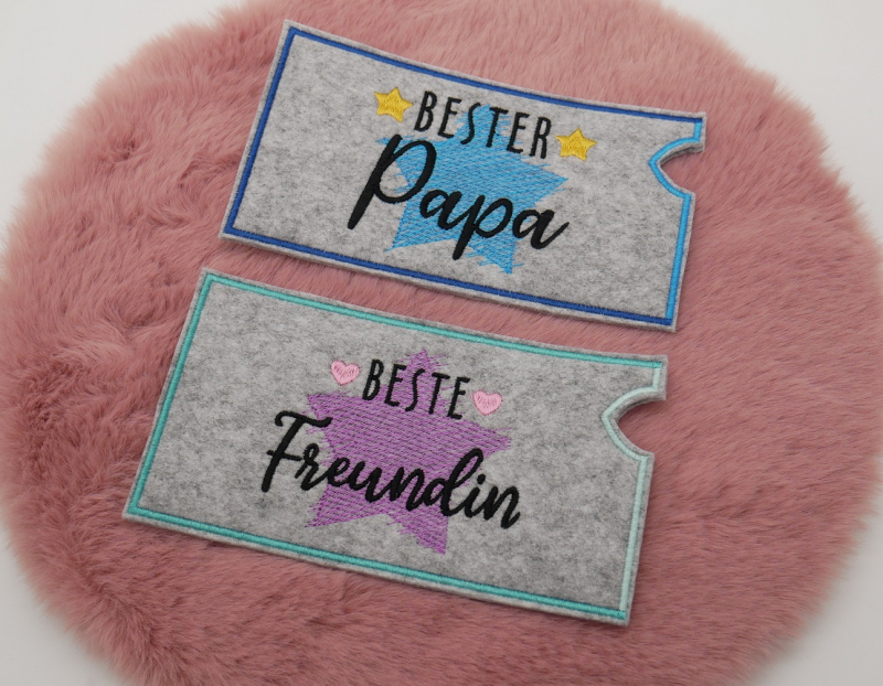 Stickdatei Set "die Besten" mit ITH Mug Rugs, Schokohüllen & Schriftzügen, Foto zeigt 2 längliche Hüllen aus grauem Filz. Einmal blauer Stern, gelbe Sternchen, Schriftzug "Bester Papa", die andere mit lila Stern, rosa Herzchen, Schriftzug "Beste Freundin"