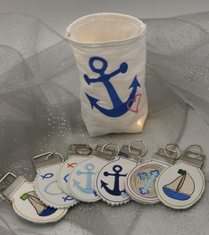 Stickdatei Set christliche Deko und Gastgeschenke (ITH), Foto zeigt hellen Lichtbeutel mit Ankeremblem, davor liegen 7 runde Anhänger. Diese mit angeklammerter Metallöse mit Schlüsselring und diversen Symbolen (Segelboot, Fische, Anker, Kreuz).