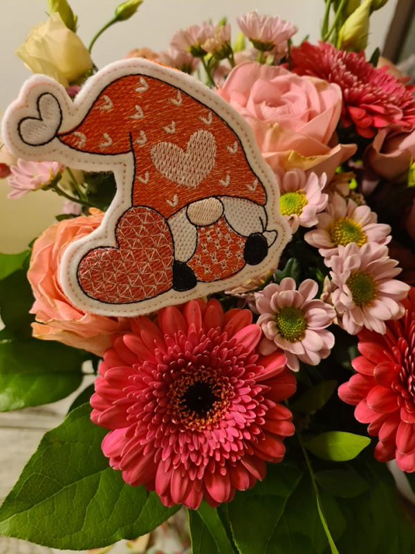 Stickdatei Set Lovegnomes ITH Stabdesigns, Foto zeigt Blumenstrauß, in dem 1 Designstab steckt. Auf hellem Untergrund Gnom mit Herz in der Hand. Rot-weiß-rosa Farbtöne. 