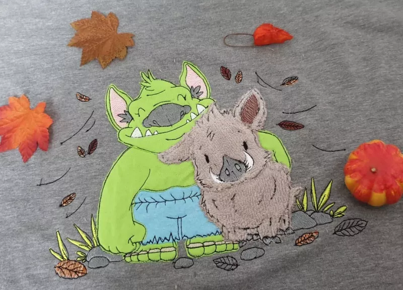 Stickdatei Trollfreundschaft Applikation (5 Größen), Foto mit Motiv auf graubraunem Stoff. Grüner Troll mit hellblauer Hose umarmt hellbraunes Wildschwein. Umgeben von gestickten Blättern, Steinen, Grasbüscheln.