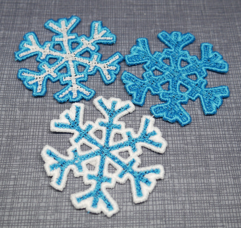Stickdatei FSL Schneeflocken-Set 6,1 x 6,3 / 9,6 x 9,9 / 17 x 17,5 cm. Foto mit 3 ausgestickten Flocken in blau-weiss, auf grauer Decke liegend. 2 diverse Strukturen.