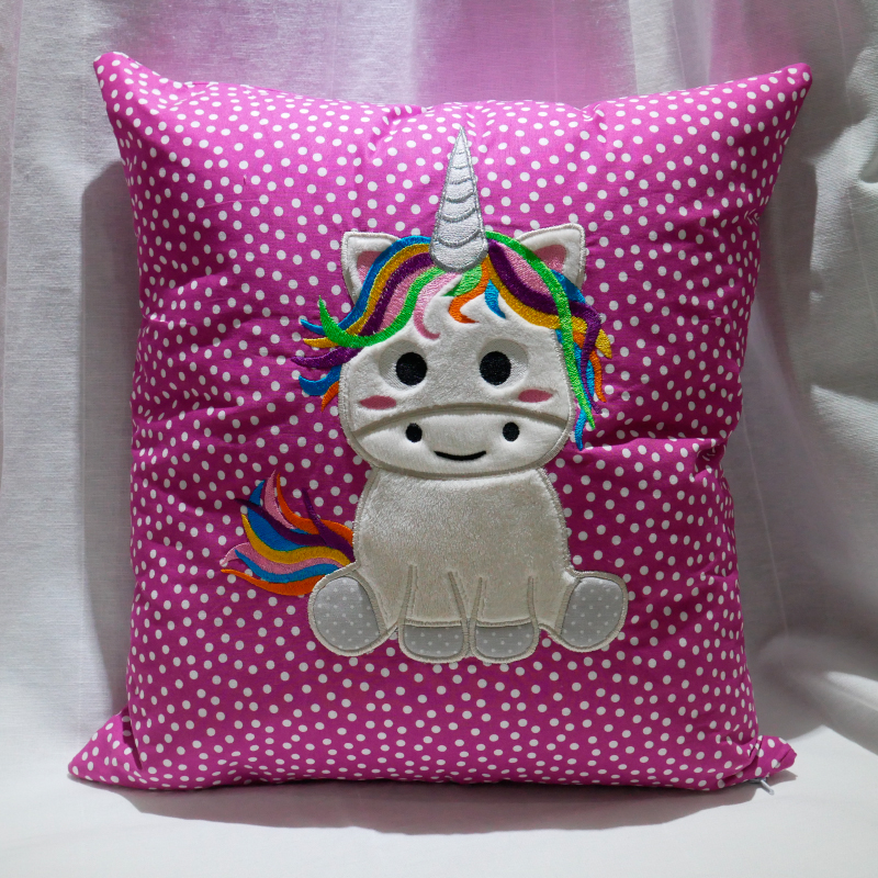 Stickdatei Sweet Unicorn (div. Rahmengrößen), Foto zeigt pink-weiß gepunktetes Kissen. Applikation sitzendes Einhorn, teilweise plüschig, bunte Mähne, bunter Schweif.