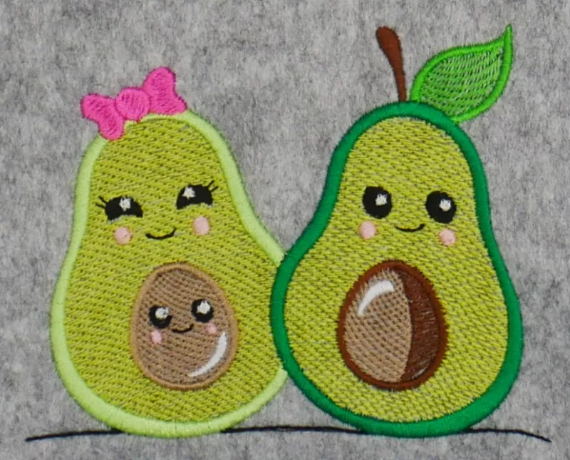 Stickdatei Set Sweet Avocados, Foto mit zwei grün gehaltene Avocados nebeneinander auf graumeliertem Filz. Die eine mit pinker Schleife und braunem Kern im Inneren, beide mit Pausbackengesicht. Die andere mit Stiel und grünem Blatt, einem Pausbackengesich