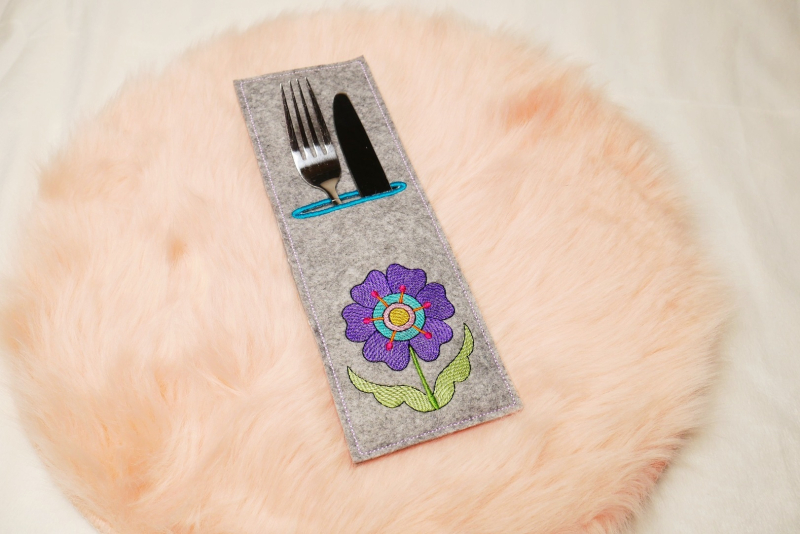 Preview: Stickdatei Set ITH Bestecktaschen Folksy Flowers (4 Größen), Foto zeigt rechteckige Hochkant-Tasche, mittelgrauer Filz, befüllt mit Besteck. Bestickung mit lila-türkis-rot-gelbem Blütenmotiv mit Stengel/Blättern in Grün.