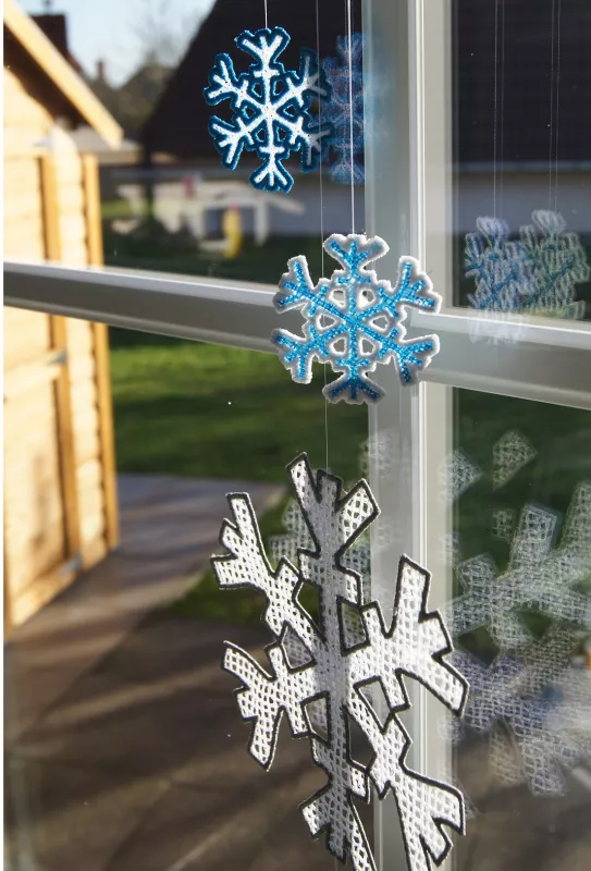 Stickdatei FSL Schneeflocken-Set 6,1 x 6,3 / 9,6 x 9,9 / 17 x 17,5 cm, Foto mit 3 Flocken unterschiedlicher Größen und Farben (vorwiegend blau und weiß). Diese hängen vor einem Fenster an je einer Schnur. Sternform, bzw. gitterartige Struktur.
