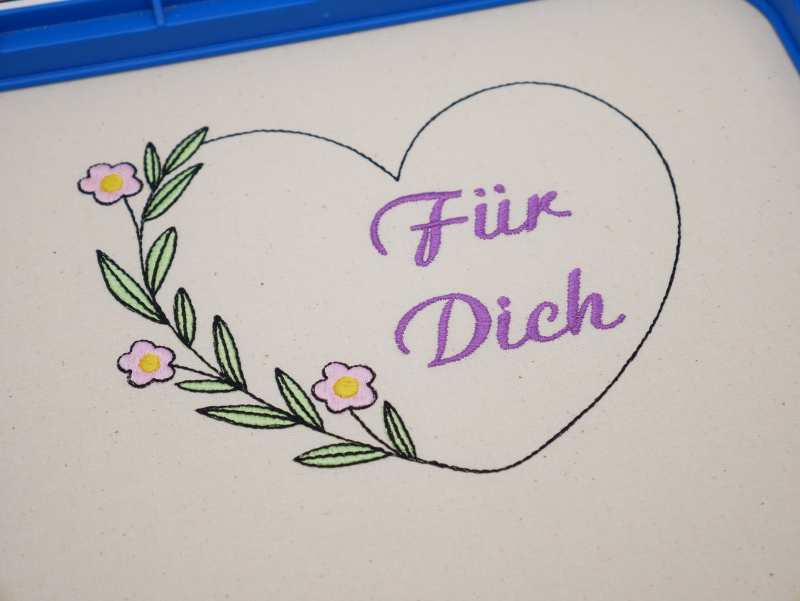 Stickdatei Set florale Rahmen + Schriftzüge, Foto mit in Stickrahmen eingespanntem hellen Stoff. Darauf Herzumriss mit Schriftzug "Für Dich" im Inneren. Herz wird auf einer Seite durch einen Blüten-Blätter-Zweig verstärkt.
