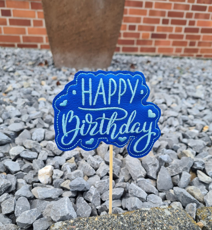 Stickdatei Set Geburtstags-Stabdesigns, Foto mit Ausfertigung, die in Kiesboden steckt, Mauer im Hintergrund. Schriftzug "HAPPY Birthday" in türkis auf mittelblauem Kunstleder, umgeben von 2 Punkten und 4 Herzen.