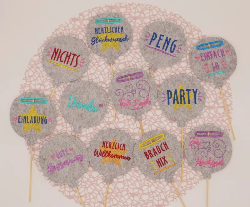 Stickdatei Set Ballon-Stabdeko ITH, Foto zeigt 12 Luftballon-Designstäbe aus mittelgrauem Filz, teilweise zusätzlich mit umrandeter Einstecköffnung. Bunte Bestickungen mit Ornamenten (Sterne, Schnörkel) und Schriftzügen ("Danke", "Nichts", "Peng"
