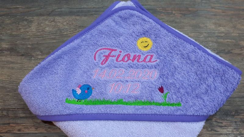 Stickdatei Set Frühlingswiese (div. Rahmengrößen), Foto zeigt bunte Bestickung auf lila Untergrund eines Babytextils. Wiese mit Blume und Vogel, Sonne, Schriftzug "Fiona 14.02.2020 10:12".