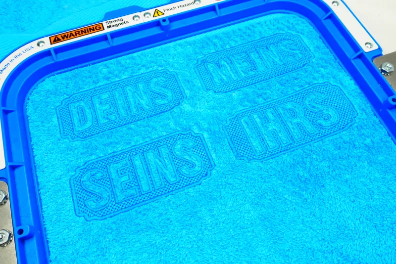 Freebie Stickdatei Set Handtuchmotive, Foto mit in Stickrahmen eingespanntem mittelblauem Frotteestoff. Vier Schriftzüge: "DEINS"/"MEINS"/"SEINS"/"IHRS", wobei die Buchstaben durch Auslassung der Bestickung entstehen, welche die Form einer länglichen Umra