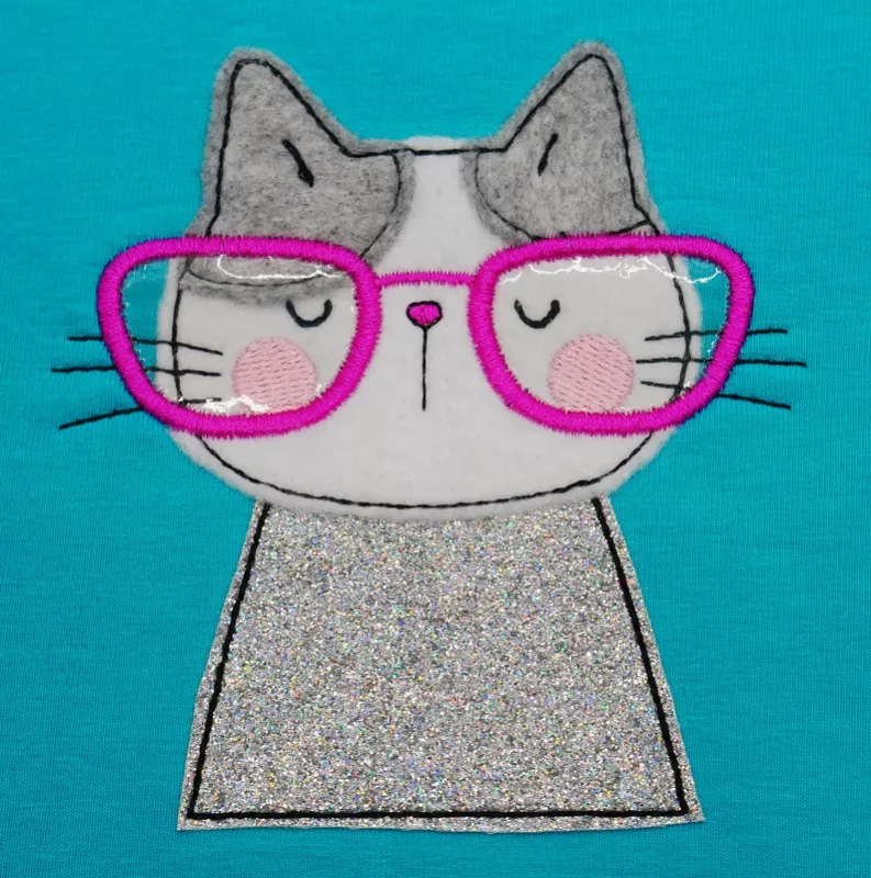 Stickdatei Set I'm Happy Cat Doodle Applikation inkl. ITH Anhänger, Foto zeigt Katzenbüste als Applikation auf türkisem Untergrund. Rosarote Brille, Katzenfarben weiß, grau und glitzerbunt.