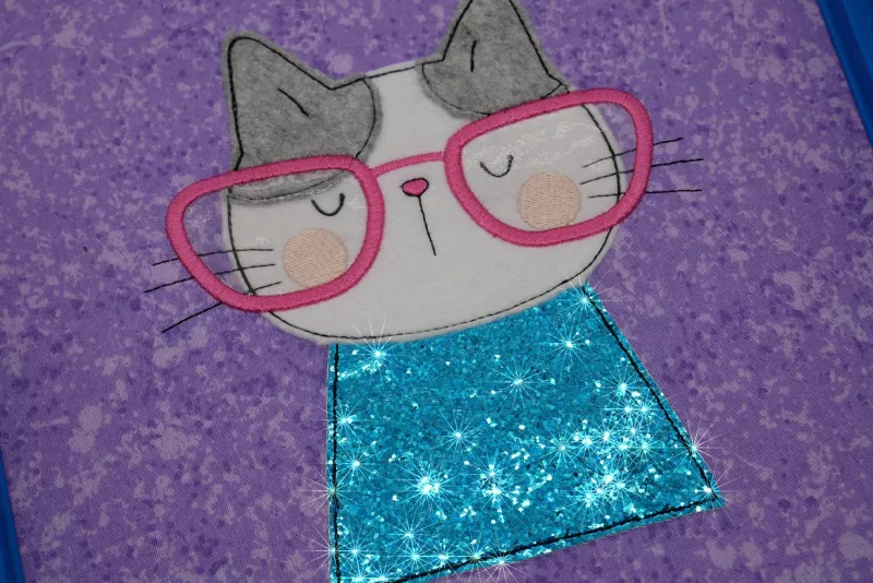 Stickdatei Set I'm Happy Cat Doodle Applikation inkl. ITH Anhänger, Foto zeigt in liegender Ansicht Katzenbüste als Applikation auf lila-gemustertem Stoff. Rosarote Brille, Katzenfarben weiß, grau und glitzerblau.