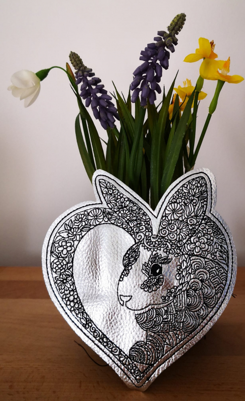 Stickdatei Set Hase mit Herz inkl. Dateien für Bestecktaschen und Stabdesign (div. Größen), Foto mit Ausfertigung vor einem Frühlingsblumenstrauß. Herz aus silbernem Kunstleder mit Bestickung in schwarz: Innenarealanteil in Musterung, daraus hervor schaut