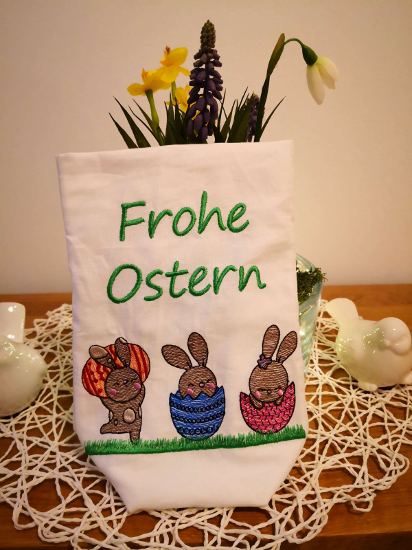 Preview: Stickdatei Set Osterhasen ITH Lichtbeutel / Utensilos, Foto zeigt hellen Hochkantbeutel vor Frühlingsdeko. Bunte Bestickung: 3 diverse Hasen mit Ei/Eierschale auf Wiese, darüber Schriftzug 
