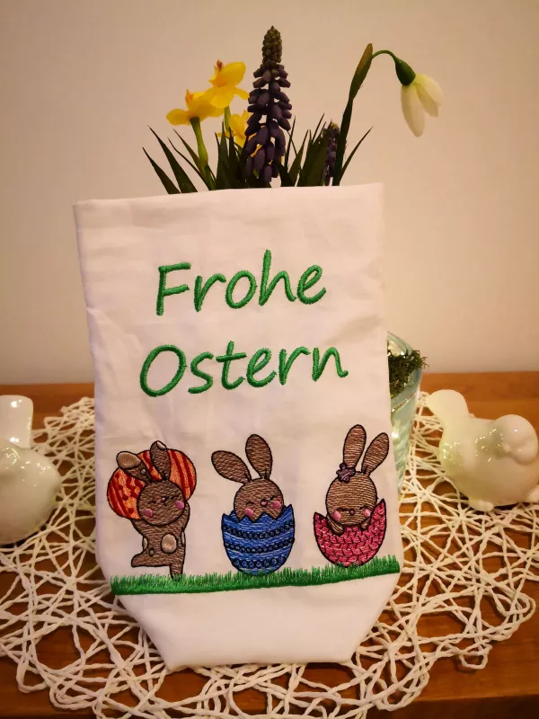 Stickdatei Set Osterhasen ITH Lichtbeutel / Utensilos, Foto zeigt hellen Hochkantbeutel vor Frühlingsdeko. Bunte Bestickung: 3 diverse Hasen mit Ei/Eierschale auf Wiese, darüber Schriftzug "Frohe Ostern".