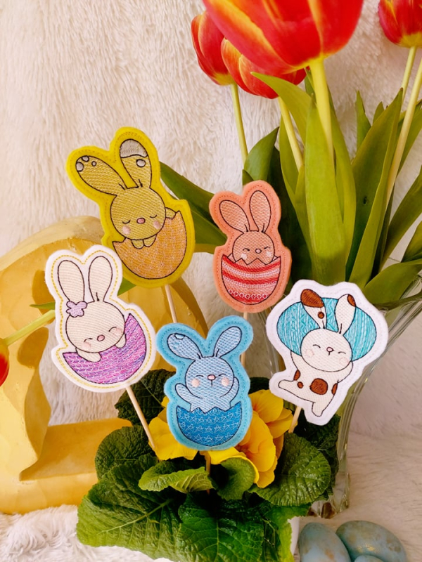 Preview: Stickdatei Set Osterhasen ITH Stabdesigns, Foto mit 5 Designstäben, in einem Blumentopf mit Blume steckend, Tulpenstrauß im Hintergrund. Motive: 4 mal Osterhase in halber Eierschale; 1 mal Osterhase, der ein Osterei stemmt.