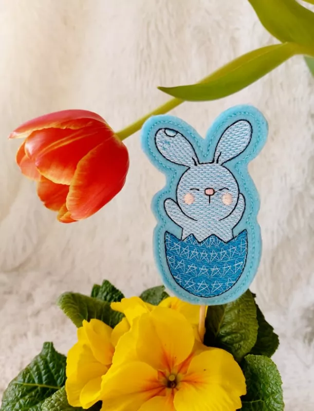 Stickdatei Set Osterhasen ITH Stabdesigns, Foto mit Designstab, in einem Blumentopf mit Blume steckend, Tulpe über dem Artikel hängend. Auf türkisem Filz gestickter Osterhase in halber Eierschale, der die Pfoten nach oben streckt.