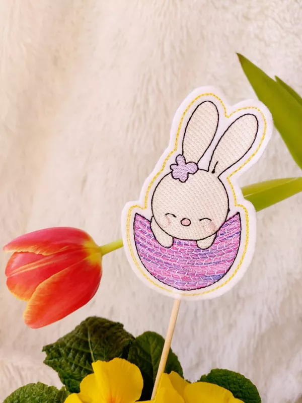 Stickdatei Set Osterhasen ITH Stabdesigns, Foto mit Designstab, der in Blumentopf mit Blume steckt, Tulpe neigt sich im Hintergrund herab. Osterhase auf hellem Filz gestickt und in halber rosa-lila Eierschale sitzend.