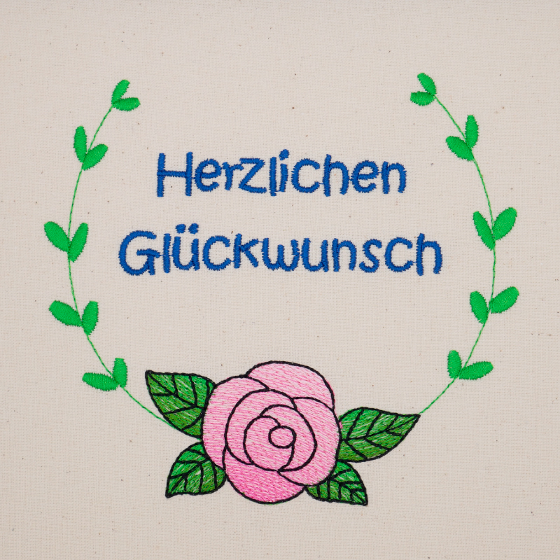 Stickdatei Set florale Rahmen + Schriftzüge, Foto mit Bestickung in grün-rosa-blau auf hellem Stoff. Von einer Rose mit Blättern führen 2 gebogene Blattzweige randgebend nach oben, dort Rahmen offen. Innen der Schriftzug "Herzlichen Glückwunsch".