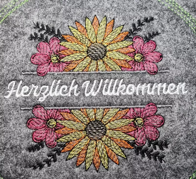 Stickdatei Set Floral Borders (div. Größen), Foto mit Ausfertigung auf grauem Filz. Arrangement mit Sonnenblume in gelb-orange-pink, ober- und unterhalb des Schriftzugplatzes identisch. Schriftzug in weiß "Herzlich Willkommen".