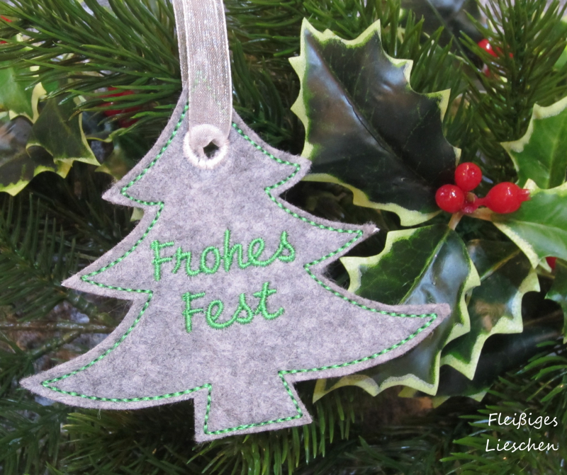 Stickdatei Set ITH Anhänger Frohes Fest, Foto zeigt 1 geösten Anhänger aus grauem Filz am Bändchen hängend, Hintergrund Weihnachtsdeko. Schriftzug "Frohes Fest", Form: Tannenbaum.