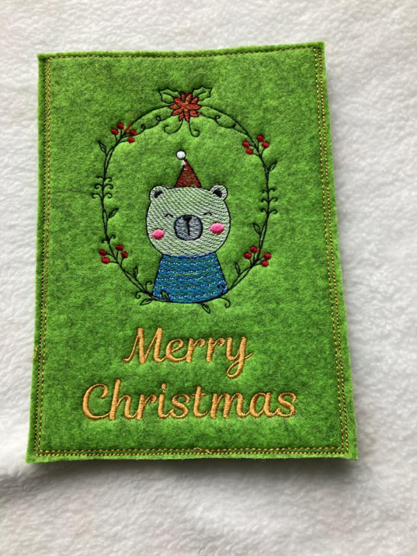 Stickdatei Set ITH Christmas-Cards - Grußkartenhüllen, Foto zeigt Hochkant-Karte aus grünem Filz. Bunte Bestickung mit Zierkranz, darin Bärchen, unten Schriftzug "Merry Christmas".