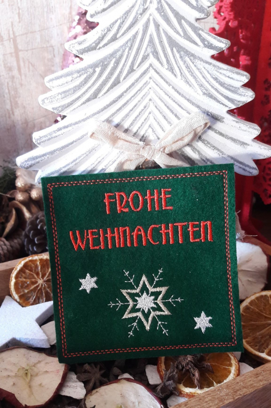 Stickdatei Set ITH Weihnachtskarten, Geschenkanhänger und Schriftzüge, Foto zeigt eine Karte vor Weihnachtsdekoration. Karte aus grünem Filz, quadratisch, Bestickung: Sterne/Eiskristall, Schriftzug "Frohe Weihnachten".