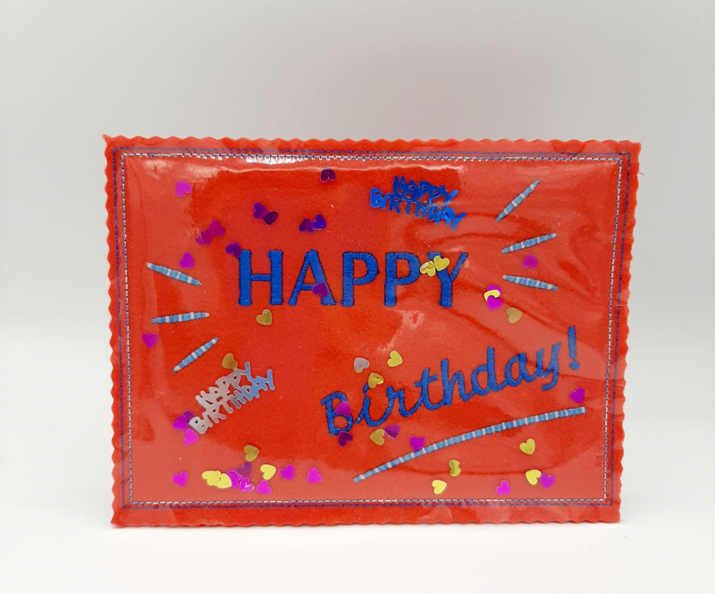 Stickdatei Set ITH Geburtstagskarten (10x10 u. 13x18), Foto mit querformatiger Geburtstagskarte in glänzendem Orange vor hellem Hintergrund. Transparente Folie auf Filz aufgesteppt bildet Zwischenraum mit eingelegten Metallstanzereien (Herzchen/Schriftzug